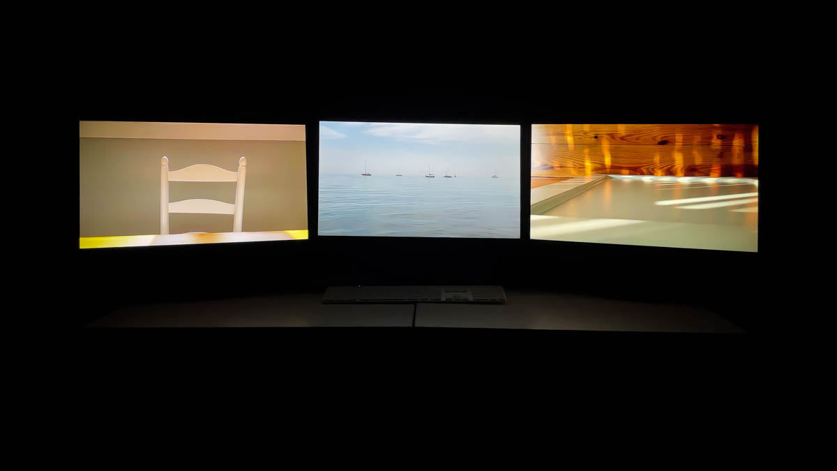 Three monitors showing the seaside magic video art by luke kurtis are the only source of light in the room.