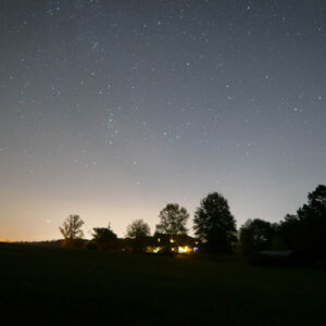 Night time photo of a house surrounded by trees with lights coming from the windows but everything around it is dark and stars fill the sky