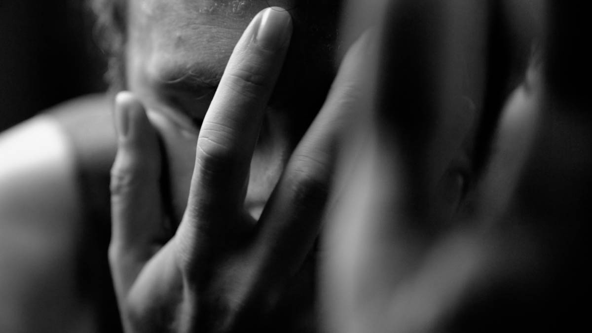 A close-up, black-and-white image of a person holding their head in their hands, eyes closed, with a distressed or contemplative expression.