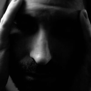 A close-up, black-and-white image of a person holding their head in their hands, eyes closed, with a distressed or contemplative expression.