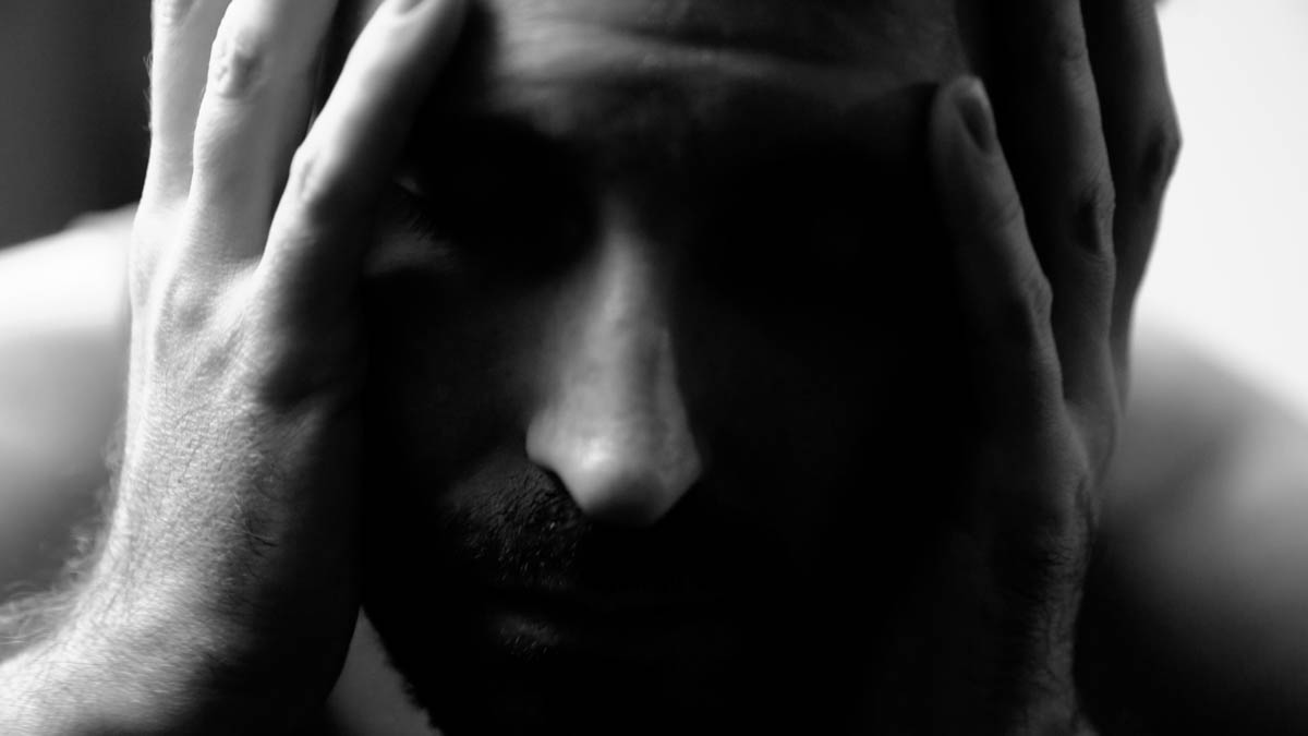 A close-up, black-and-white image of a person holding their head in their hands, eyes closed, with a distressed or contemplative expression.