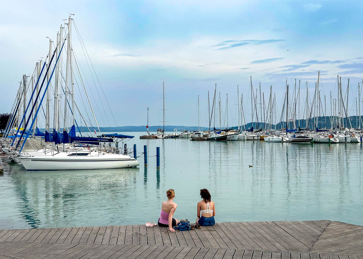 Two women sit on a wooden dock by a calm marina, facing the water with many sailboats anchored. The sky is partly cloudy, and the overall atmosphere is peaceful and serene.