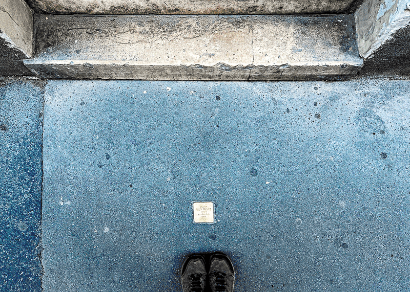 A pair of shoes stands on a worn blue-gray sidewalk facing a stone doorstep. A small commemorative plaque is embedded in the pavement near the shoes. / A person stands on a stone path facing the edge of a riverbank, where old shoes, some filled with flowers and yellow ribbons, are arranged as a memorial. The water of the river is visible beyond the edge.