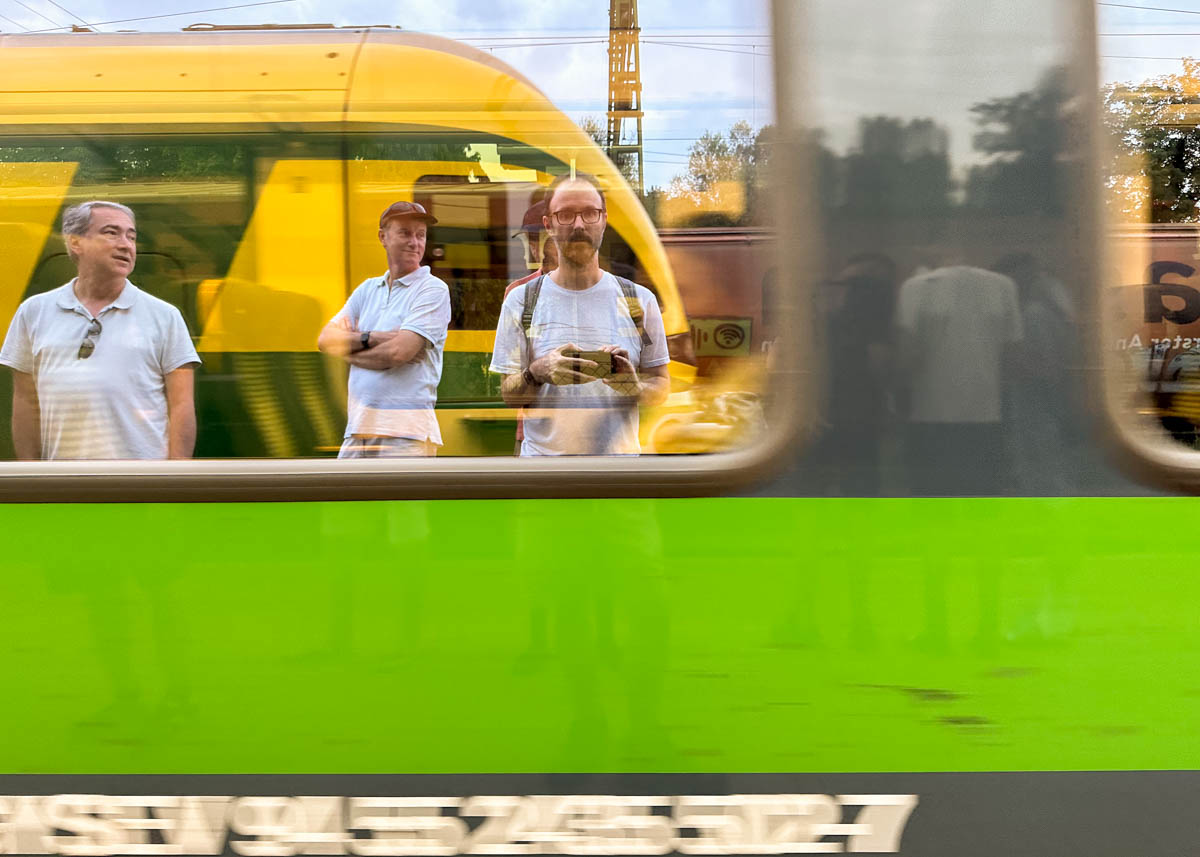 Three men in white shirts are reflected in the window of a passing green and yellow train, with blurred motion partially obscuring their images. A yellow train and other background details are visible through the window.