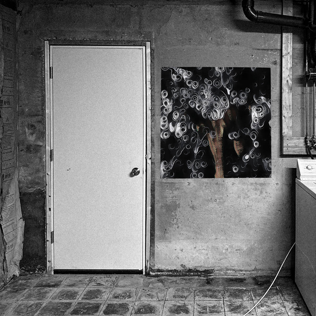 Black-and-white basement interior with a white door beside a poster of the original ‘a harras’ album cover, whose subtle color contrasts with the otherwise monochrome image.