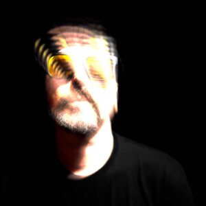High-contrast, front-facing portrait of a man in a black beanie and sunglasses against a pitch-black background. His face is heavily blown out with white light, overlaid with a distorted, double-exposed projection of warm orange horizontal scan lines across his eyes and forehead.